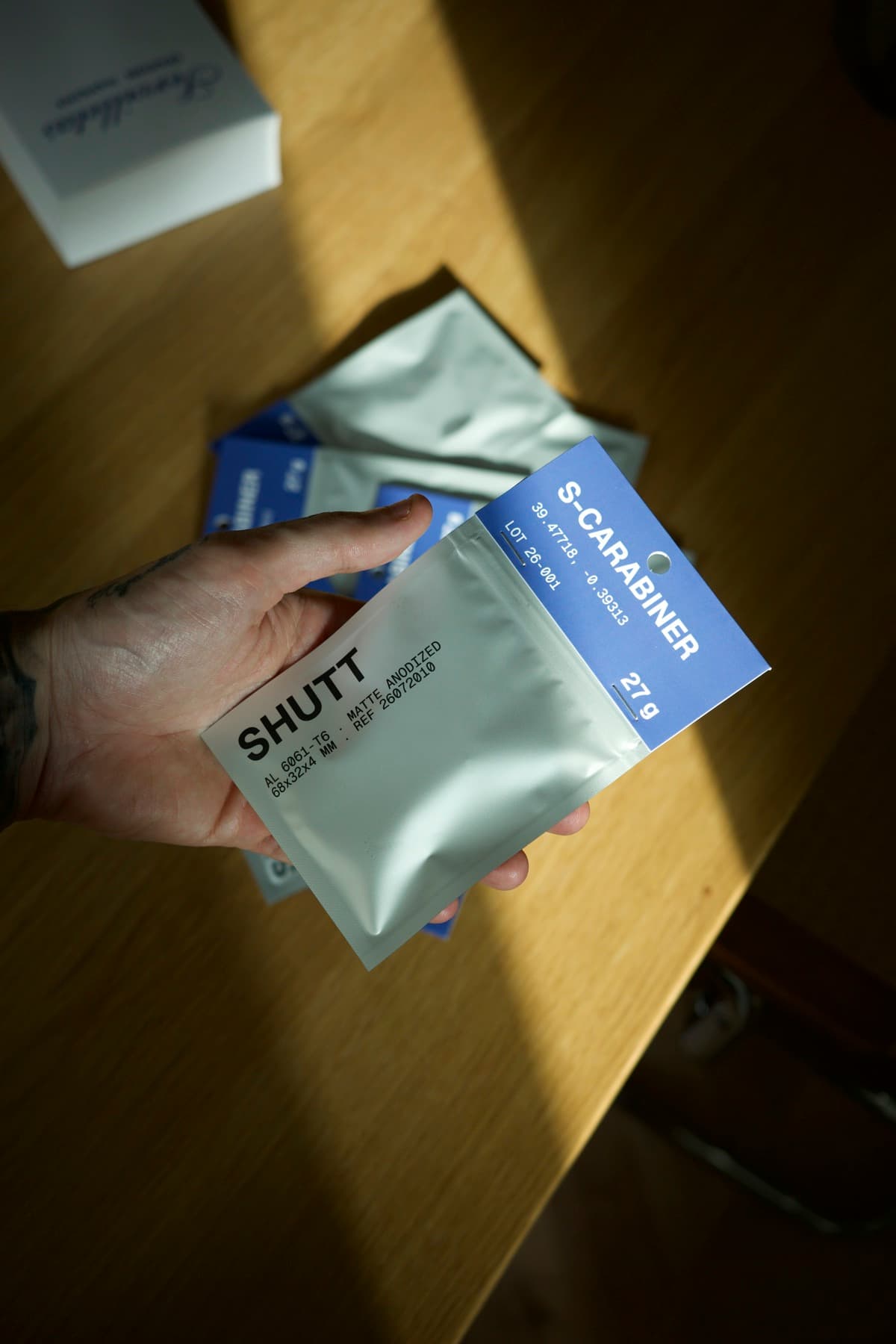 S-CARABINER in its foil pouch with the blue header card, held over a wood table with more pouches behind